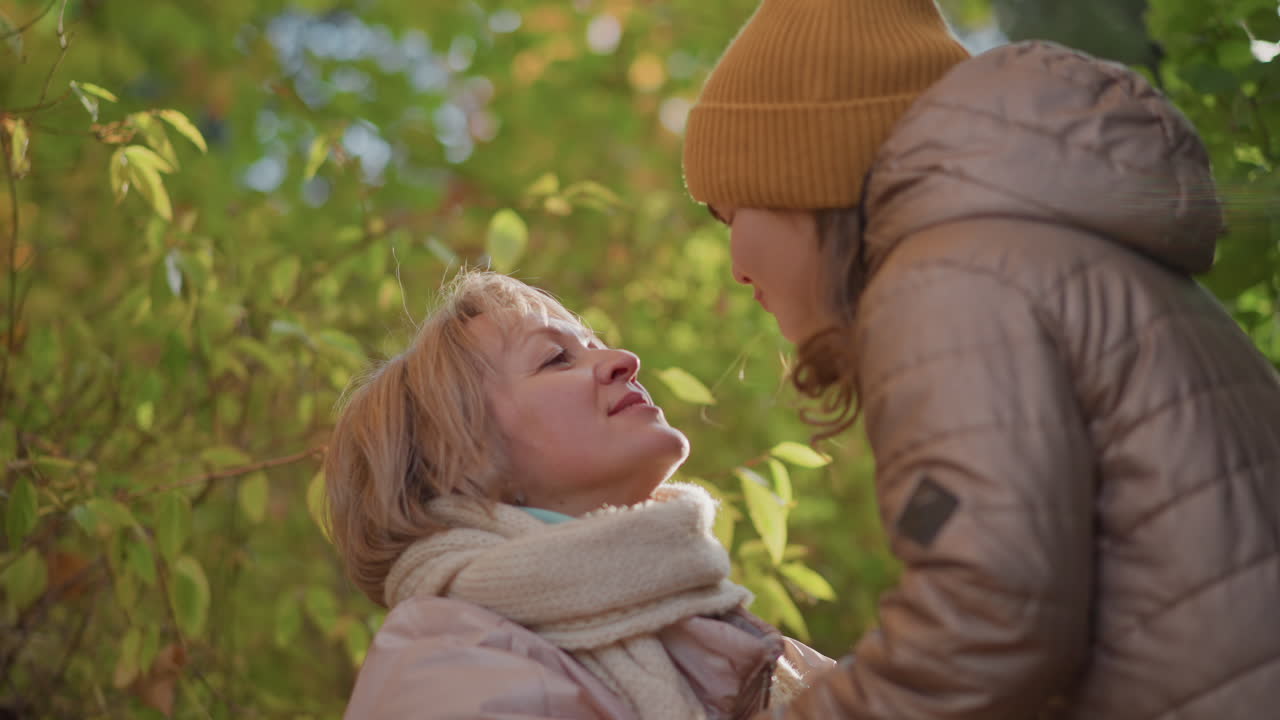 kid kisses mom on lips, playfully pushing mom backward as she smiles, warm autumn light filters through colourful foliage, both in jackets and knit hat, capturing joyful playful family moment