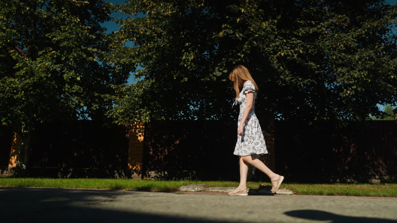 Side view of elegant young woman walking alone along sunny suburban street, showcasing floral gown with grace and confidence under clear blue sky and soft shadow of tree leaves