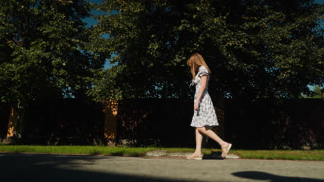 side view of elegant young woman walking alone along sunny suburban street, showcasing floral gown with grace and confidence under clear blue sky and soft shadow of tree leaves