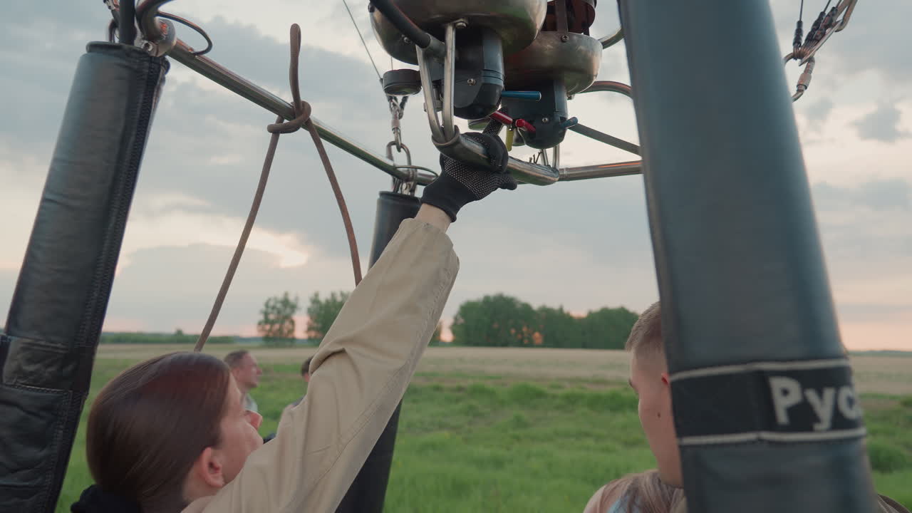 woman in glove reaches up to burner control increasing flame into colorful hot air balloon envelope over green field at sunset with wicker basket and rope tethers for launch preparation