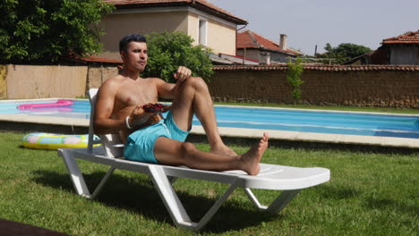 young fit man eats cherry near villa poolside, static view