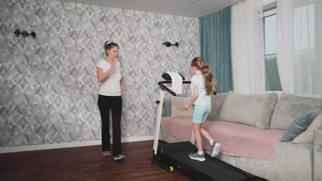 little girl runs on mid-speed treadmill while female trainer stands nearby offering encouragement inside bright living room with curtain-draped windows, gray wall pattern, and plush couch