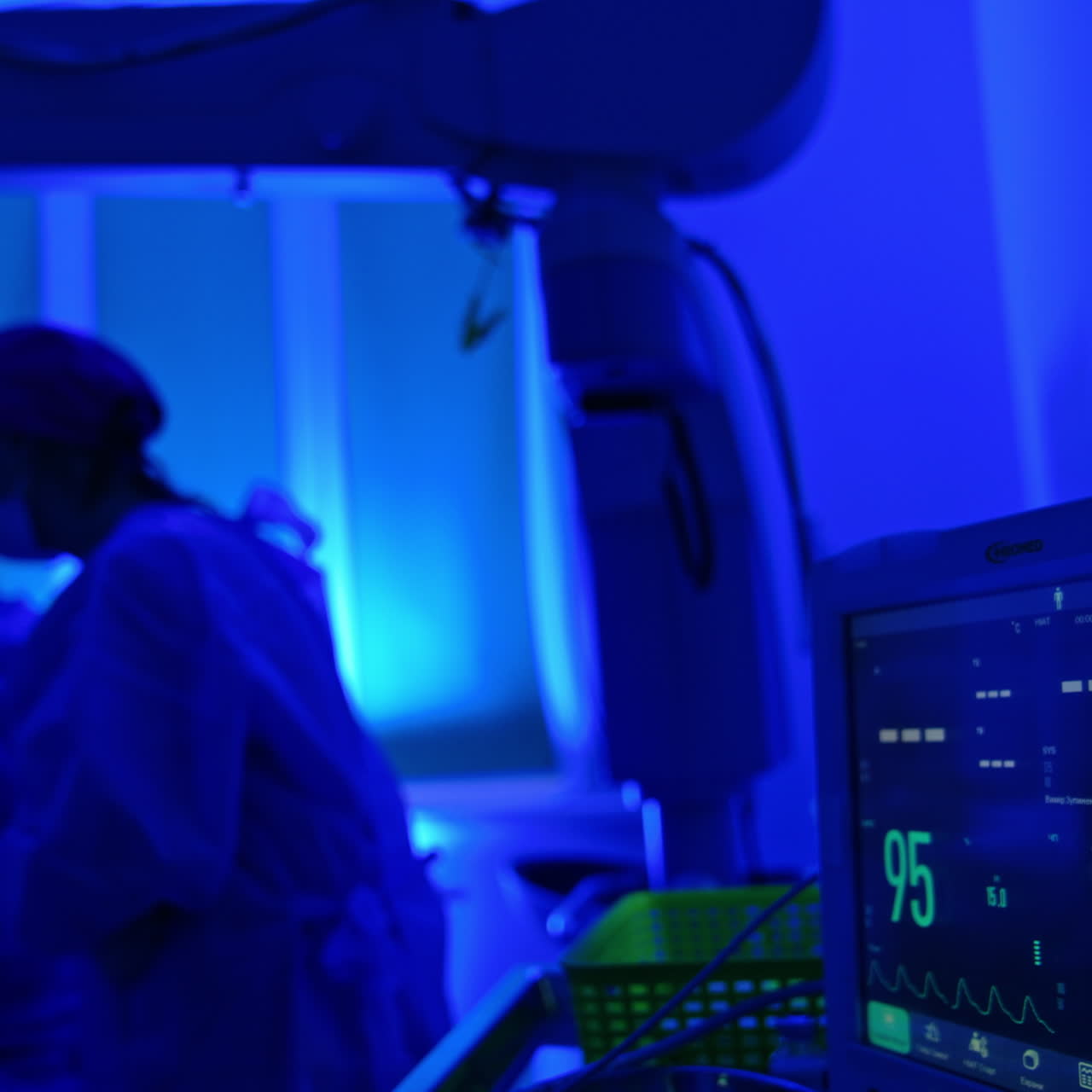 Screen with patient's life signs is in the surgery room with blue light. Two doctors sit at microscope at backdrop in blur