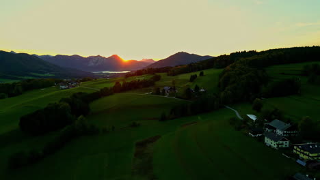 aerial drone backward moving shot over green farmlands along mountain slope in norway with sun rising in the background