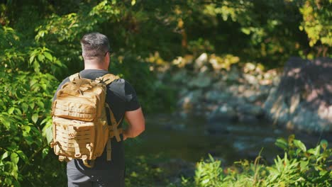 Male-hiker-arriving-a-mountain-river-in-slow-motion-surounded-by-boulders-and-trees-during-hike-in-jungle