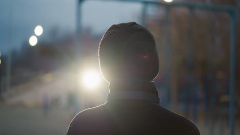 back view of a person in a black outfit and beanie, lifting a ball up with a headset around their neck, the background features a blur of city lights and a reflective glow in the distance