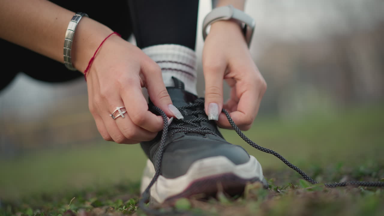 Close up das mãos amarrando os cadarços do tênis antes do treino ao ar livre. Anéis e smartwatch visíveis, amarração e aperto cuidadosos na sola texturizada, meias brancas, plano de fundo com parque gramado, preparação enérgica