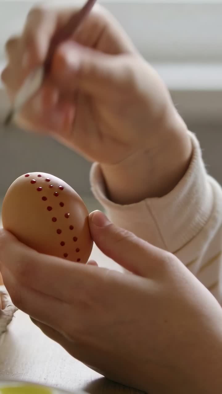Vertical video: Holding egg to show holes, hands painting red dots to decorate by window with brush