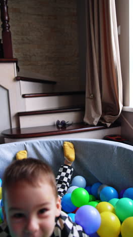 toddler in checkered pajama jumps into the dry pool. smiling kid lies on the colorful balls looking at camera.