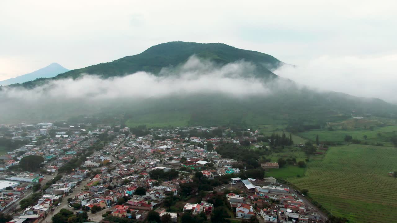 Aerial: cityscape and cloudy mountain top over Tuxpan during the day in Jalisco, Mexico, pull out drone shot