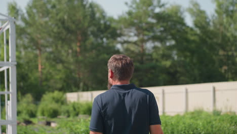 behind view of man in blue polo walking up farm path through green rows of plants past white metal frame structure under bright sun, rural garden scene with tool belt and gentle breeze over leaves
