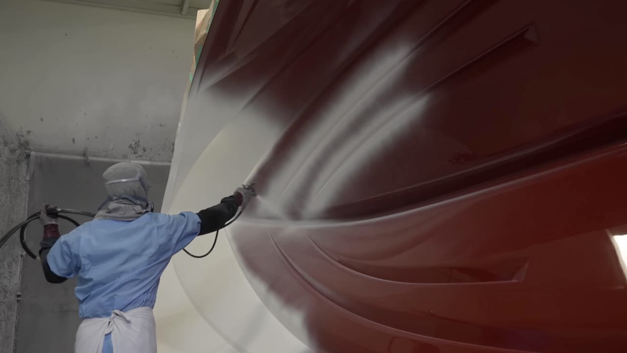 Peintre professionnel en combinaison de protection utilisant un pistolet de pulvérisation pour appliquer une sous-couche blanche sur la coque d'un grand bateau rouge dans un chantier naval