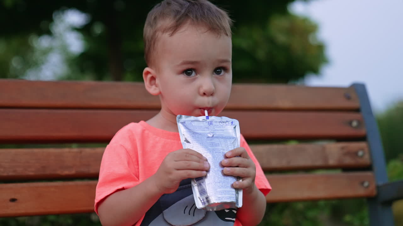 Lovely dark-eyed Caucasian toddler sits on the bench outdoors. Baby boy drink juice from a straw and pack.