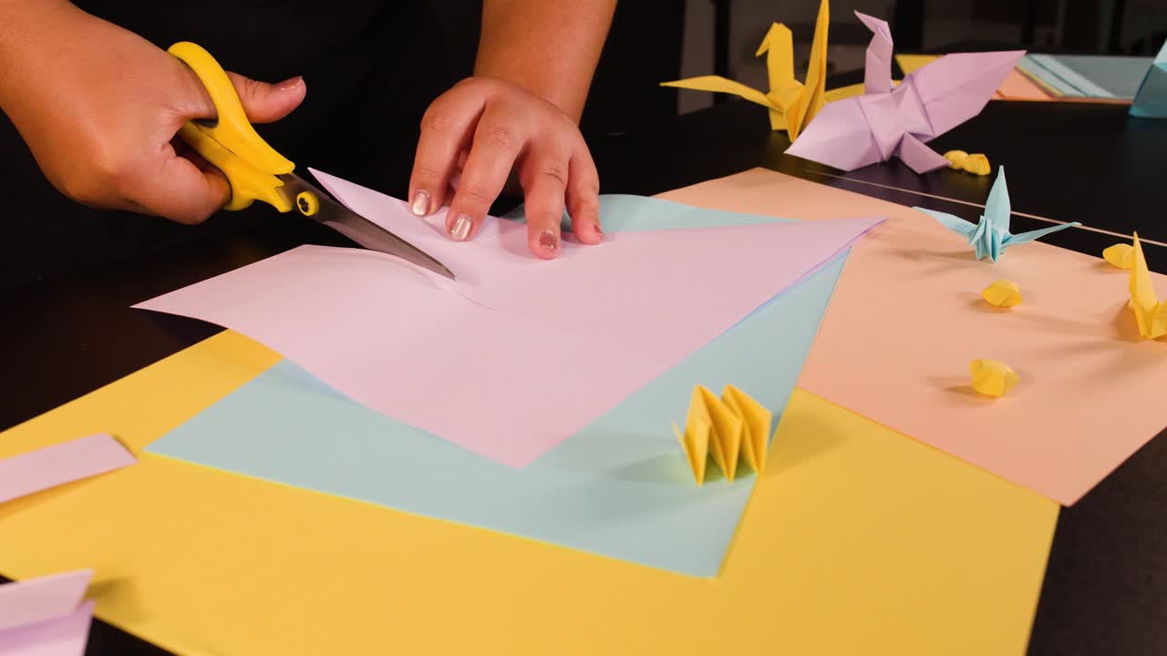 A person uses yellow scissors to cut pink paper on a desk with origami cranes