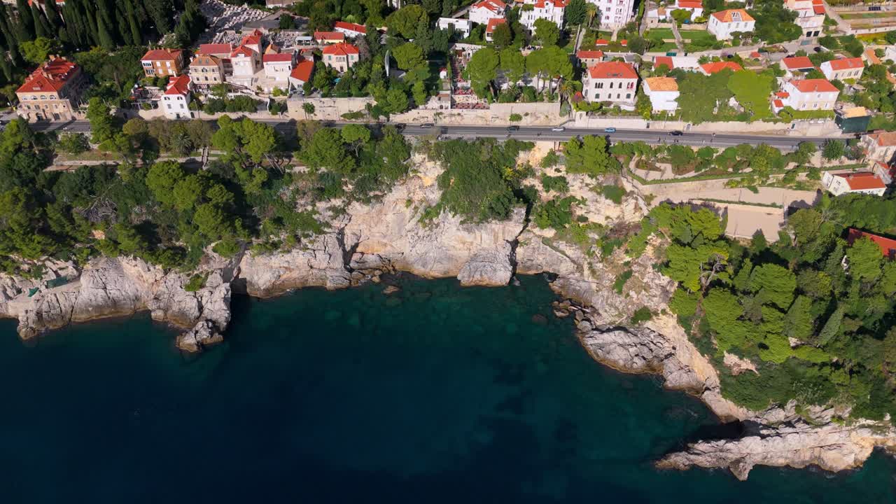 High-angle aerial drone captures Dubrovnik’s cliffside coast, with a narrow road hugging the edge and cars passing by, terracotta-roofed buildings below, and the deep blue Adriatic Sea