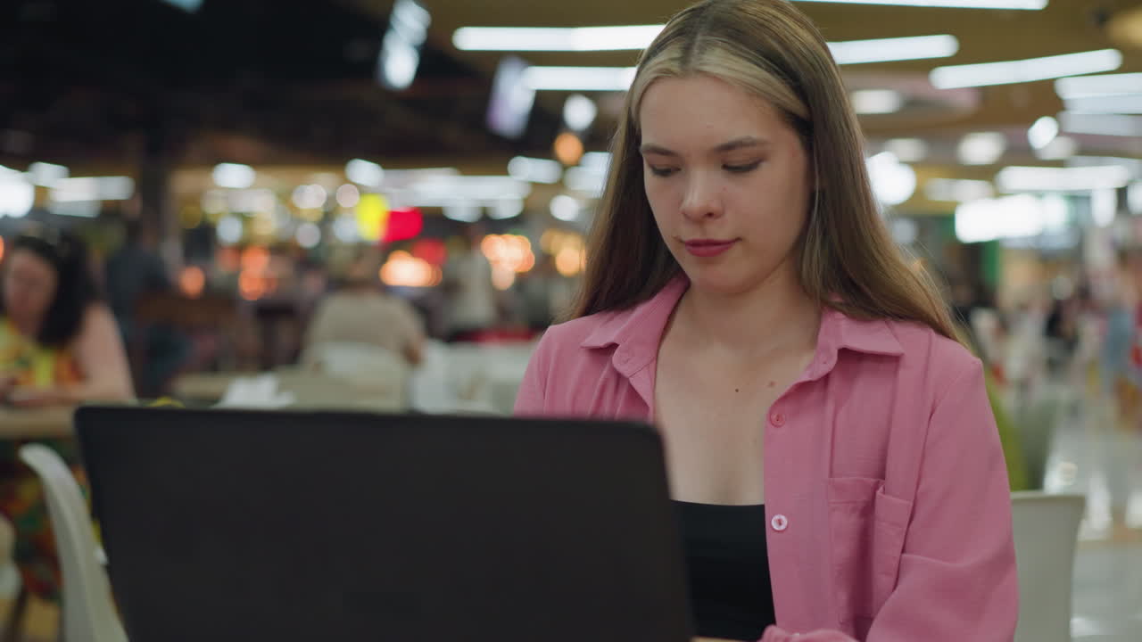 mujer joven en un vestido rosa sentada en un restaurante trabajando en su portátil mientras se mueve ligeramente en una diversión, movimiento de baile con una sonrisa suave, fondo borroso de personas comiendo con luz borrosa