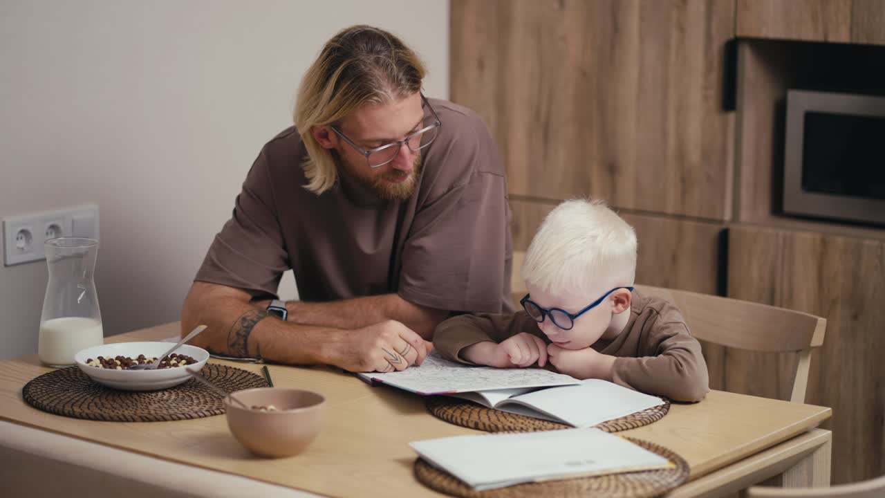 feliz niño albino con cabello blanco en gafas azules redondas se le enseña a leer y escribir con la ayuda de libros de texto especiales en la escuela y su padre rubio le ayuda con la tarea en una cocina moderna