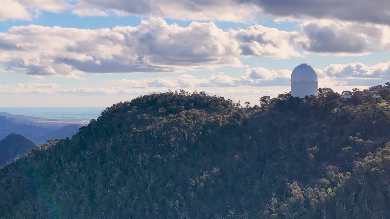 Drone footage captures a white astronomical observatory dome atop a lush, forested mountain ridge