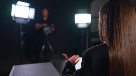 unrecognized brunette lady sits her back to camera talking and gesturing with her hand. cameraman can be seen at blurred backdrop working backstage.