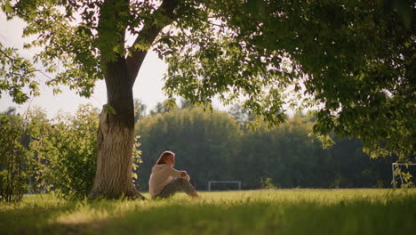 lady seated under tree with arms folded over her legs, in thoughtful reflection, gently moving leaves create a tranquil atmosphere, with background of green field and distant goalpost