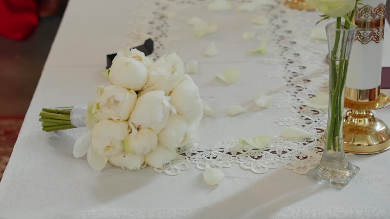 Elegant wedding ceremony table decorated with white rose bouquet, lace cloth, gold candleholder, and scattered petals creating a romantic and sacred atmosphere