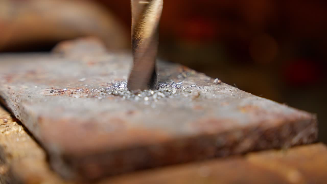 Hand-held drill penetrates flat metal sheet on wooden base, bits of metal scatter outward