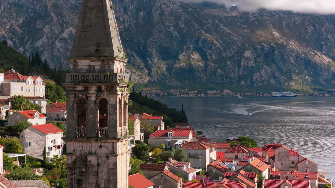 Pemandangan udara dari drone menara lonceng di Perast yang menjulang di atas rumah-rumah batu tradisional, dengan Teluk Kotor dan pegunungan curam terlihat di belakangnya. Teluk Kotor, Montenegro