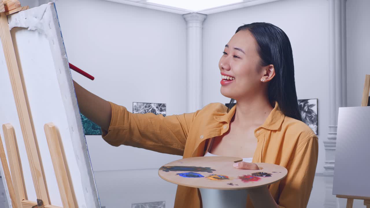미소 짓는 아시아 여성 예술가가 갤러리에서 오일 페인트와 으로 캔버스에 행복하게 그림을 그리고 있습니다.