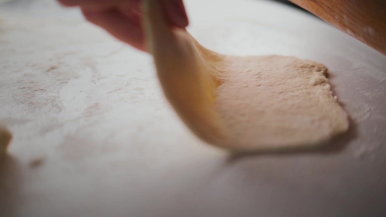 Hand uses a wooden roller to flatten dough on a floured surface. Fingers guide the tool with gentle pressure, ensuring even thickness. Focused, patient work in a warm, homely kitchen