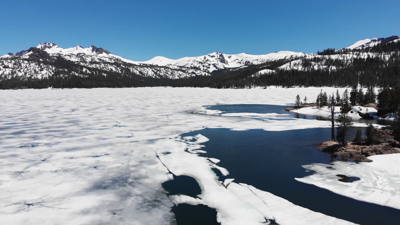 Ilmakuva jäätyneestä järvestä kevään sulamisaikaan, taustalla mäntymetsä ja lumihuippuiset Sierra Nevadan vuoret