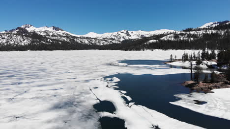 Toma-Aérea-Sobre-Un-Lago-Congelado-Durante-El-Deshielo-De-Primavera,-Bosque-De-Pinos-Con-Montañas-Nevadas-De-Sierra-Nevada-En-El-Fondo