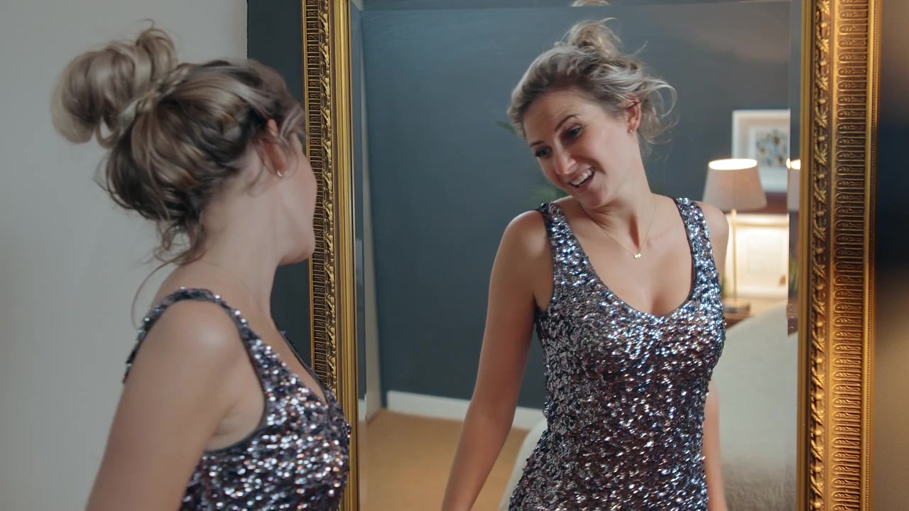 Giovane donna che prova un vestito in preparazione per una grande serata, guardando il suo riflesso nello specchio, ballando ma non essendo felice.