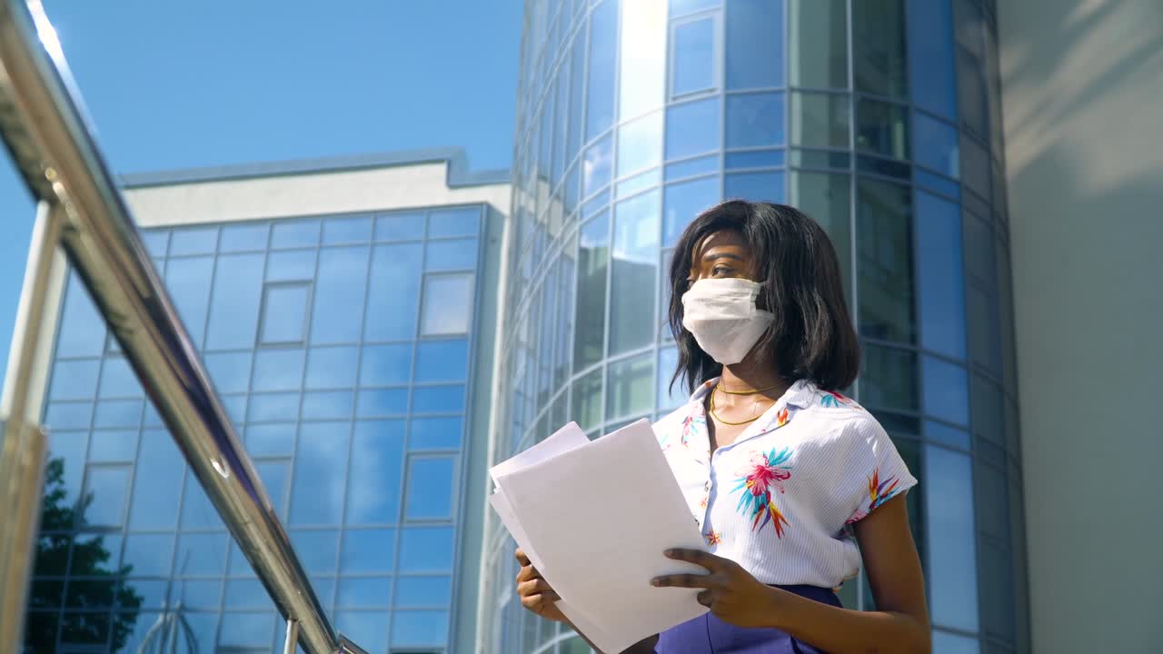 African american young businesswoman with documents in a protective mask looking at the camera near modern office building. End of quarantine