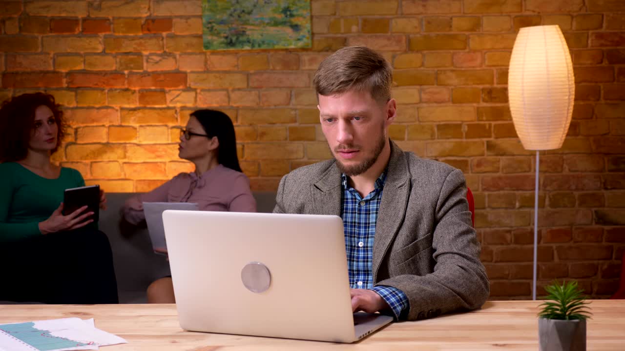 Closeup shoot of adult businessman using the laptop looking at camera and smiling indoors in the office. Female employee discussing data with a colleague on the background
