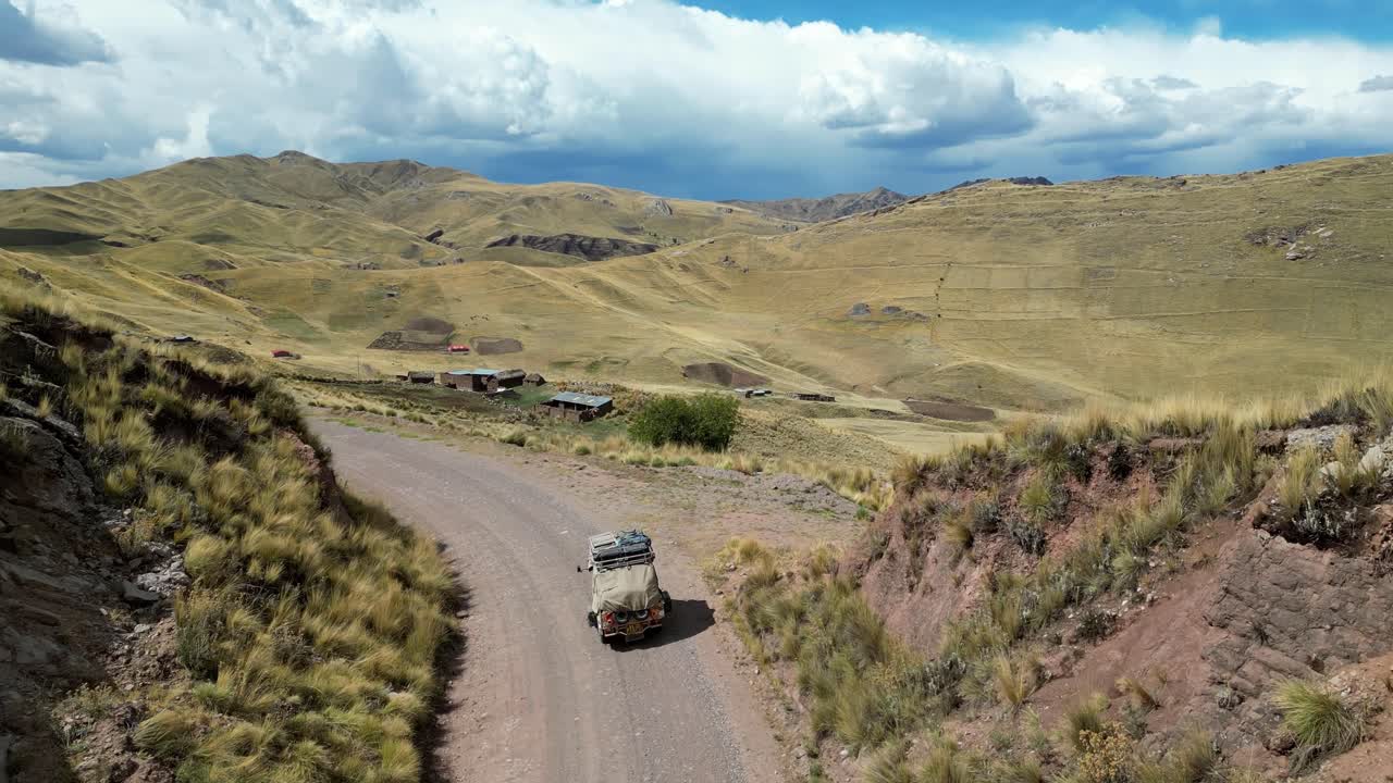 4K aerial view of a tuk tuk crossing grassy Andean plains, blades of grass rippling in the breeze as the vehicle moves through golden light