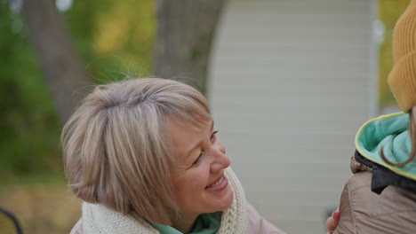 granny collecting yellow leaf from daughter hand then gently pulling daughter close and pecking cheek on autumn park bench surrounded by golden foliage and falling leaves