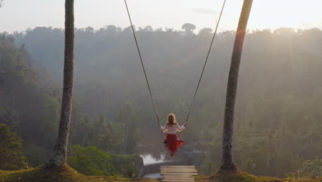 aerial-view-woman-swinging-over-tropical-rainforest-at-sunrise-sitting-on-swing-with-view-of-river-enjoying-having-fun-on-holiday-travel-freedom