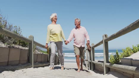 Happy-senior-caucasian-couple-holding-shoes-in-their-hands-walking-on-path-leading-to-the-beach