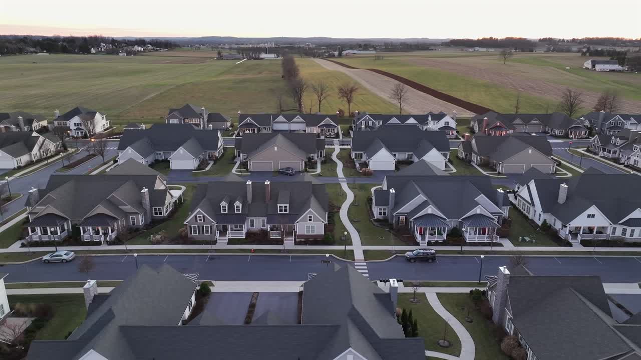 Modern semi-detached houses with grey roof in new developing area of America town. Rural farm fields and countryside in distance. Aerial lateral wide shot. Cold winter sunset in USA.