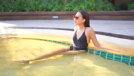 An-attractive-young-woman-lounging-on-the-steps-leading-into-a-resort-pool-tilts-her-face-toward-the-sun