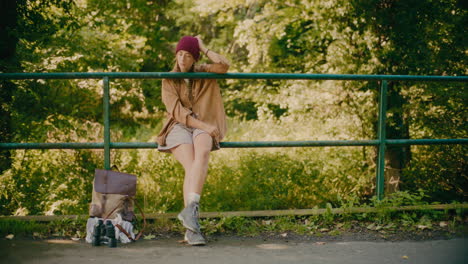 sad young woman sitting on railing by backpack in forest