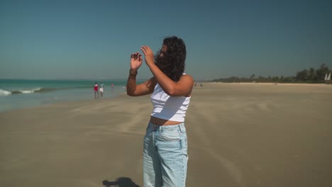 Brunette-girl-putting-her-hair-up-in-a-white-t-shirt-on-a-beach-with-other-people-walking-by