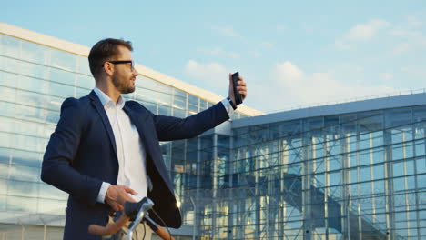 Handsome-man-wearing-glasses-and-suit-is-taking-seifie-pictures-on-the-smartphone-at-a-big-airport-or-other-city-building