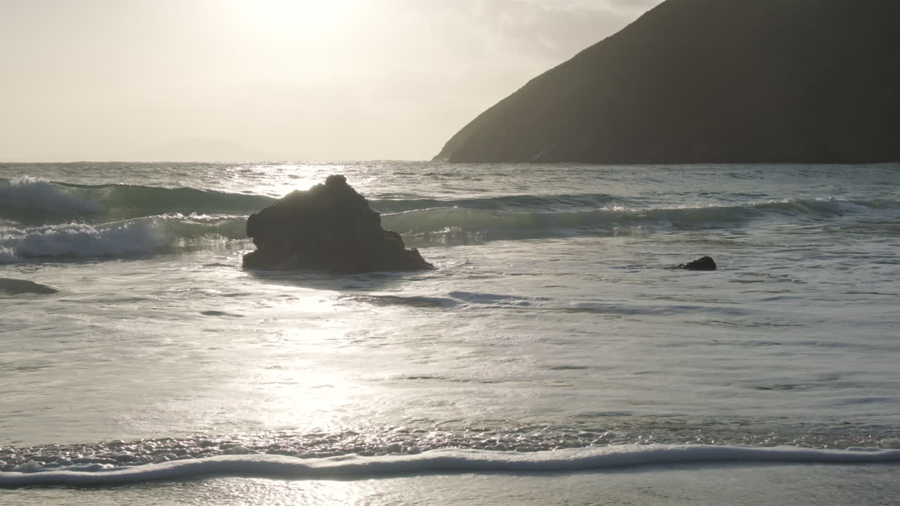 Wide shot of a single stone in the ocean. Huge hill in the background. Waves hitting the stone in slow motion. Silhouette shot with backlight from the sun.