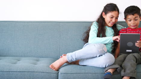 brother and sister sitting on sofa at home using digital tablet