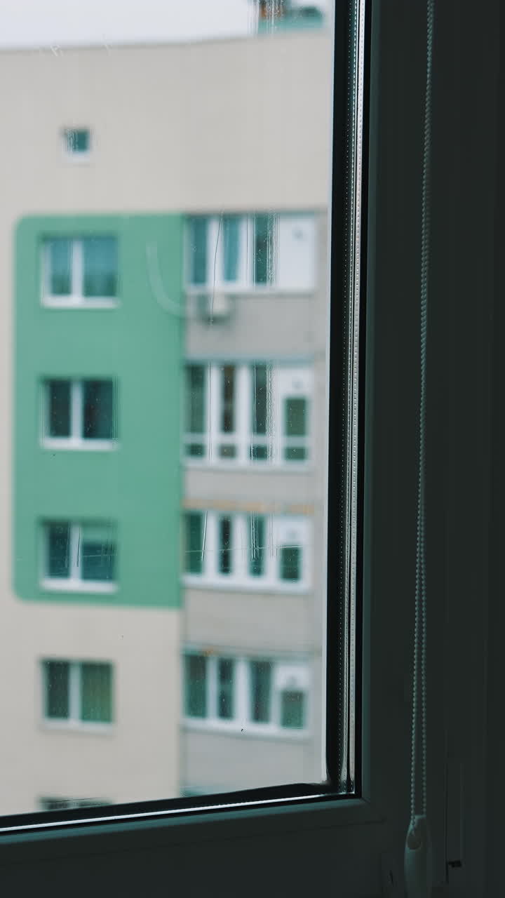 Man cleans the window and squeezes water with a brush. Male worker washing glass inside the flat. Block of flats background through clean window. Householding chores. Vertical video