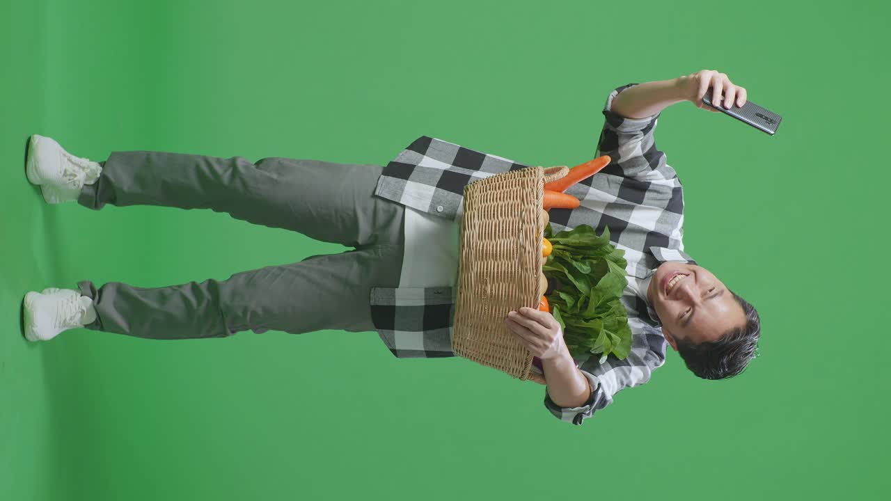 cuerpo lleno de granjero masculino asiático con canasta de verduras tomando selfie con teléfono inteligente en el fondo de la pantalla verde en el estudio