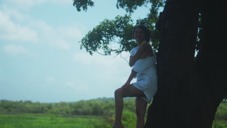 barefoot woman with long curly 3c hair and warm brown skin poses wrapped in a white dress against a wide tree trunk, surrounded by lush tropical greenery and soft daylight in a serene setting