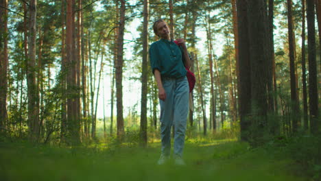 woman walking through serene sunlit forest with red backpack slung over shoulder, adjusting strap with calm expression, surrounded by tall trees and lush greenery