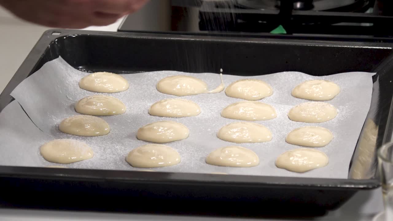 A person prepares macaron batter by piping small discs onto parchment paper. The process takes place in a contemporary kitchen with a clean, organized workspace
